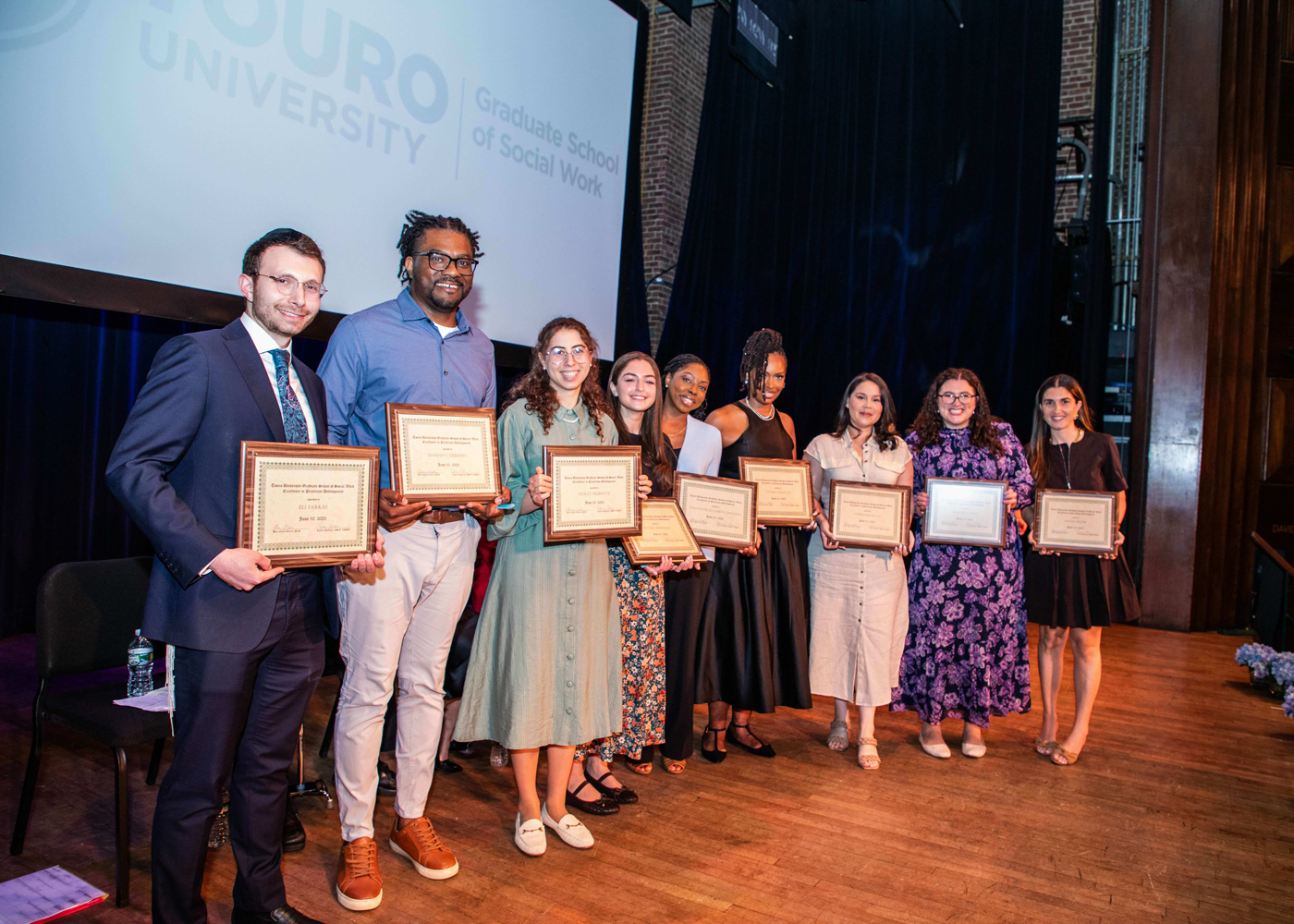 Nine students smile while standing on a stage holding award plaques