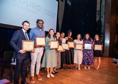 Nine students smile while standing on a stage holding award plaques