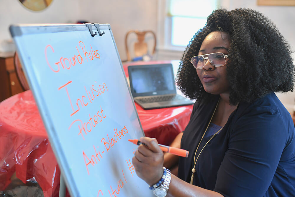 Professor Shakira Kennedy writing in orange marker at an easel whiteboard. Words you can see are Ground Rules, Inclusion, Protest, Anti-Blackness