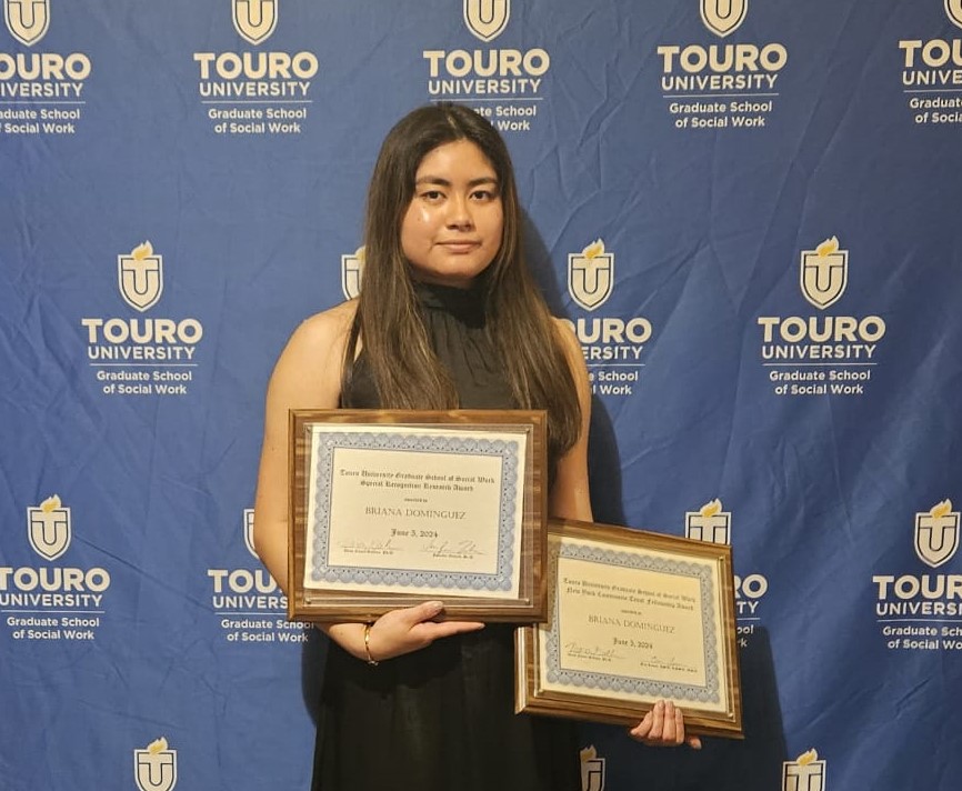 female student standing facing camera against Touro backdrop holding two framed awards