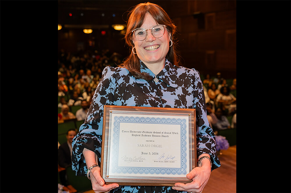 Sarah Orgel standing and smiling facing camera holding a framed award in front of her