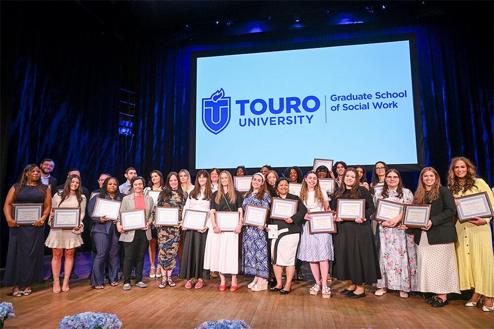 Dozens of young women lined up on a concert hall stage smiling at the audience while holding framed certificates of their awards for their accomplishments.