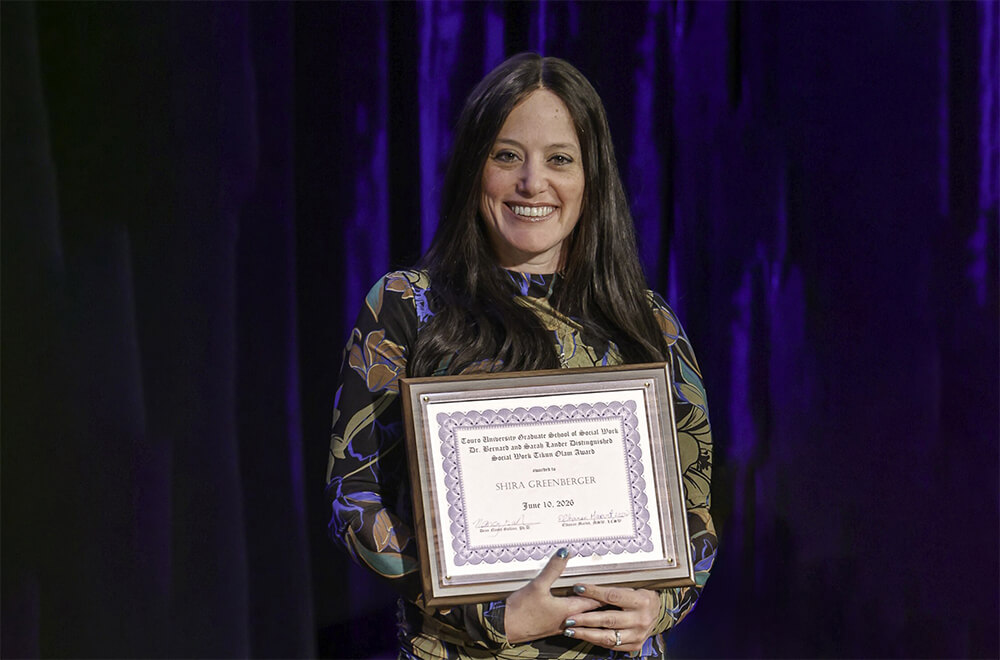 Woman holding framed award and smiling at audience from stage. 